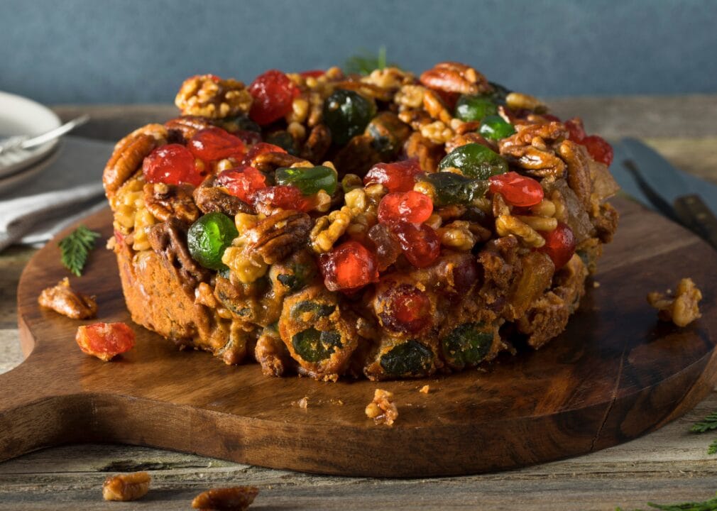 Fruitcake A round brown cake on a wooden cutting board with sprinkled nuts and candied fruit.