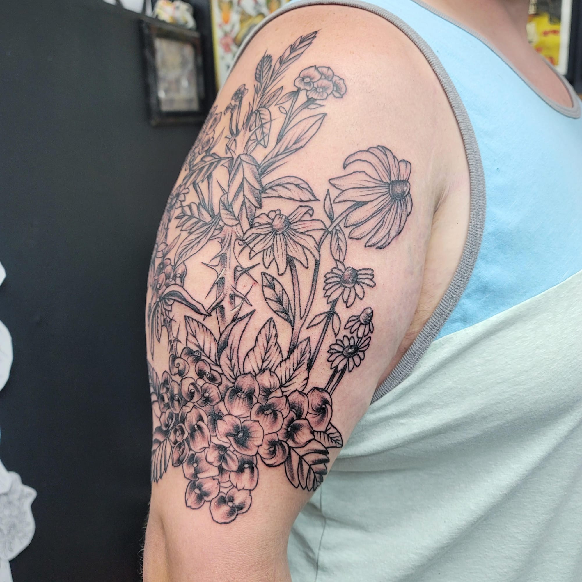 Botanical elegance blooms across this upper arm in an exquisite black-and-grey wildflower garden that captures nature's delicate beauty with stunning linework precision. The artist masterfully balanced various flower species - from bold echinacea blooms to clustered pansies and tall wheat stalks - creating a cohesive meadow scene that flows naturally with the arm's contours. This sophisticated piece proves that monochromatic floral tattoos can be just as captivating as their colorful counterparts 🌾