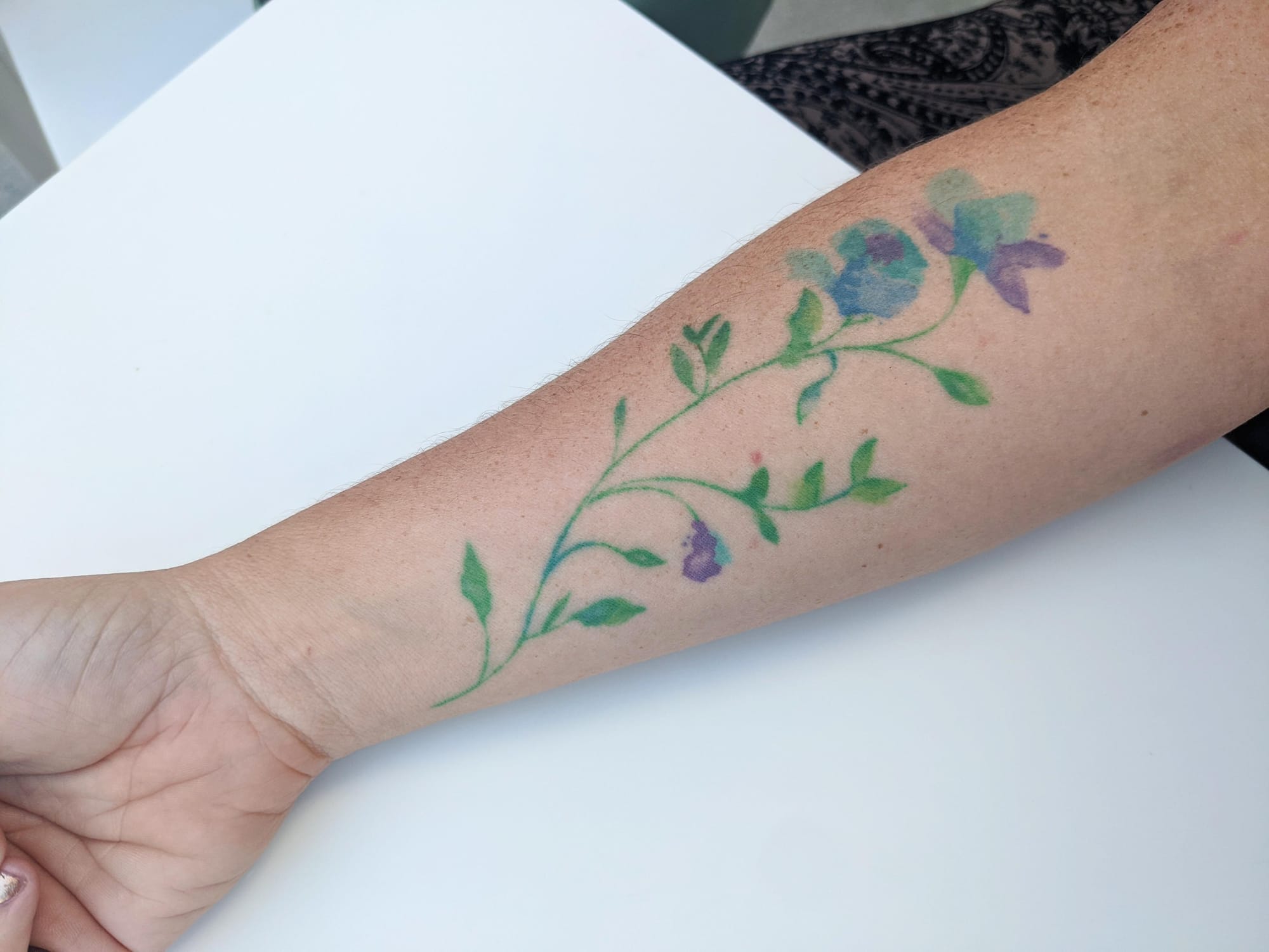 Delicate watercolor blooms cascade down this forearm in dreamy blues and purples, connected by graceful green vines that seem to dance across the skin. The soft, painterly technique creates an ethereal garden that looks like it was brushed on with liquid starlight 🌸