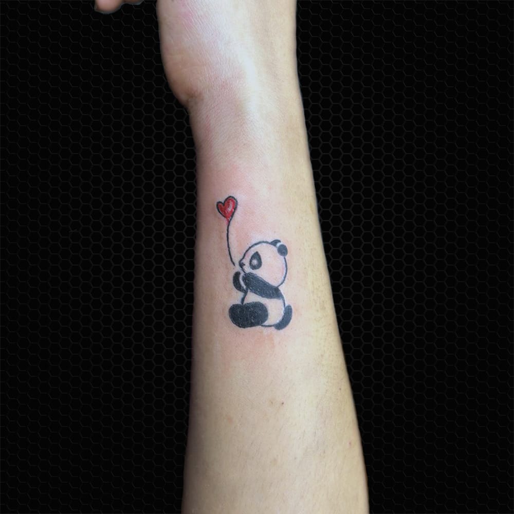 A delicate panda clutches a crimson heart balloon in this charming forearm piece that perfectly captures childhood innocence with modern minimalist flair. The simple black linework creates an almost sketch-like quality, while the single pop of red adds just enough color to make the design feel alive and whimsical. 🐼