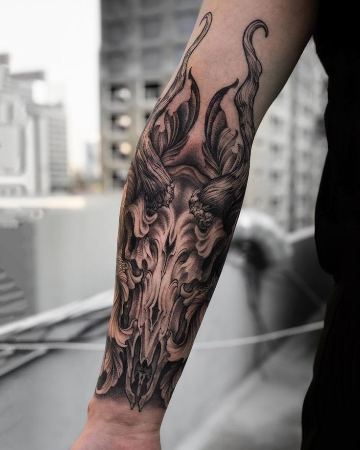 Masterful black and gray artistry transforms this forearm into a canvas of mythical power, where an intricately detailed dragon skull emerges through swirling flames and ornate baroque flourishes. The exceptional depth and dimensional shading creates an almost sculptural quality that seems to move with the contours of the arm 🔥 Every delicate line and shadow demonstrates why this piece belongs in a tattoo museum.