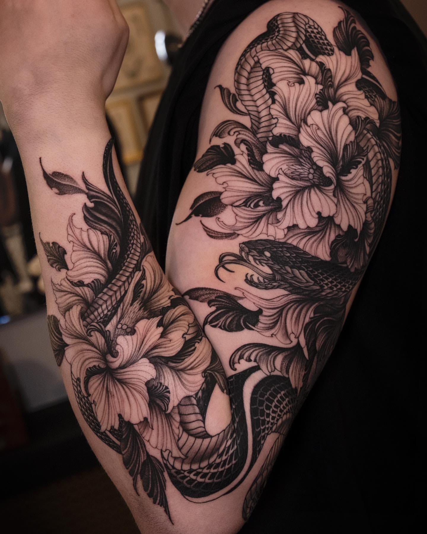 Flowing serpentine forms dance with blooming florals in this breathtaking black and grey sleeve, where traditional Japanese motifs come alive through masterful linework and shading. The artist's skillful use of negative space allows each petal and scale to breathe, creating a harmonious balance between the powerful serpents and delicate blossoms that wrap organically around the arm's natural contours. 🐍