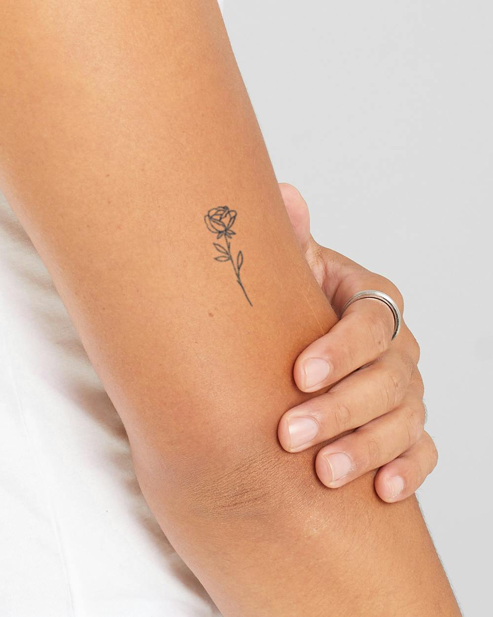 Delicate line work transforms a simple rose into pure poetry on skin, with its minimalist stem gracefully following the natural curve of the forearm. The understated black ink and precise botanical details create a timeless piece that whispers rather than shouts, perfect for those who appreciate subtle elegance. 🌹