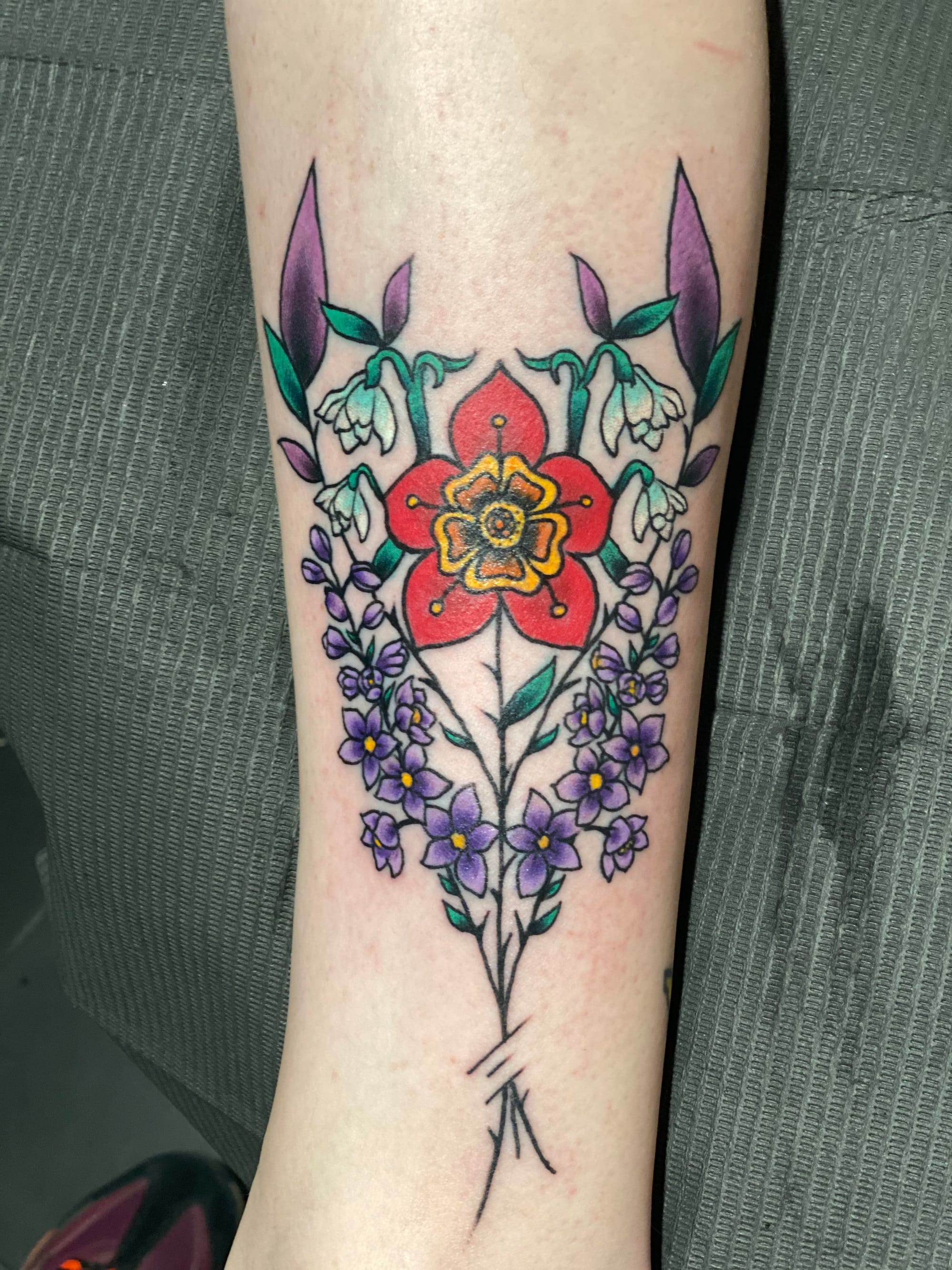 Delicate folklore meets modern artistry in this enchanting floral bouquet that transforms the forearm into a living canvas 🌸 The intricate design features a striking red bloom at its heart, surrounded by cascading purple violets and crowned with graceful white bell flowers, all tied together with flowing ribbon details. Rich traditional tattooing techniques bring depth and dimension to each petal, creating a piece that feels both timeless and deeply personal.