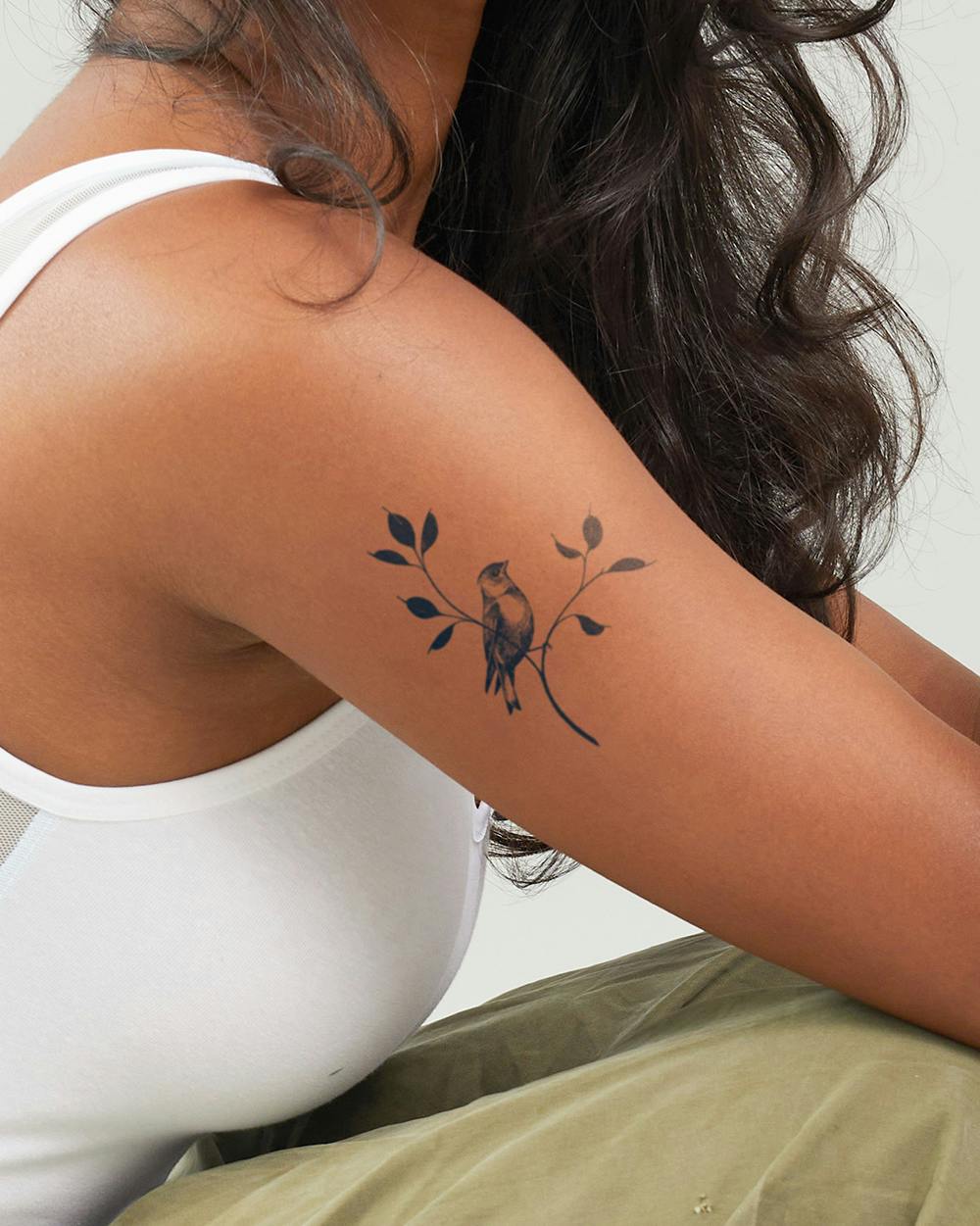 Delicate branches cradle a perched songbird in this beautifully balanced shoulder tattoo that flows naturally with the body's contours. The soft, muted tones and organic linework create an almost watercolor effect, while the placement along the upper arm allows the design to peek gracefully from tank tops and sleeveless pieces 🕊️