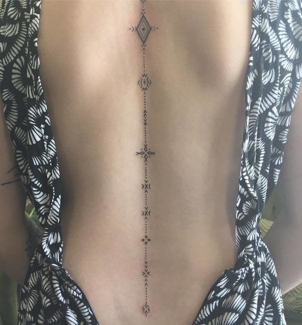 A delicate spine tattoo creates an elegant vertical journey down the back, featuring geometric diamonds and intricate dotwork that follows the natural curve of the body. The fine line work and symmetrical pattern transforms the spine into a decorative centerpiece, while the tribal-inspired elements add depth and cultural richness to this stunning placement ✨