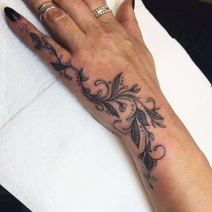 Delicate botanical artistry flows seamlessly from finger to forearm in this stunning blackwork piece, where ornate leaves and swirling flourishes create an organic sleeve effect. The intricate dotwork and flowing linework transform the hand and wrist into a canvas of natural elegance, proving that floral tattoos can be both feminine and boldly sophisticated. 🌿