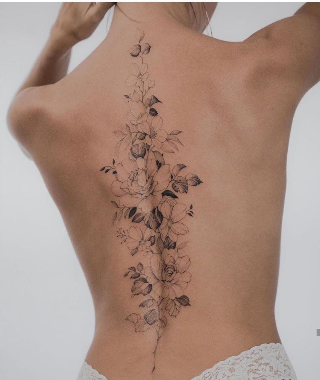A breathtaking botanical masterpiece flows down the spine like nature's own watercolor painting, featuring delicate roses, peonies, and wildflowers rendered in stunning black and grey linework. The intricate detailing and organic placement create an almost ethereal garden that seems to bloom directly from the skin, transforming the back into a living canvas of feminine grace 🌸
