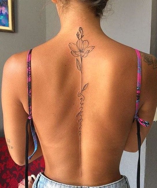 A delicate floral stem flows gracefully down the spine in this stunning line art piece, featuring an elegant bloom at the top with script lettering trailing below like falling petals. The minimalist black ink design creates beautiful symmetry along the natural curve of the back, proving that sometimes the most powerful tattoos are also the most understated ✨