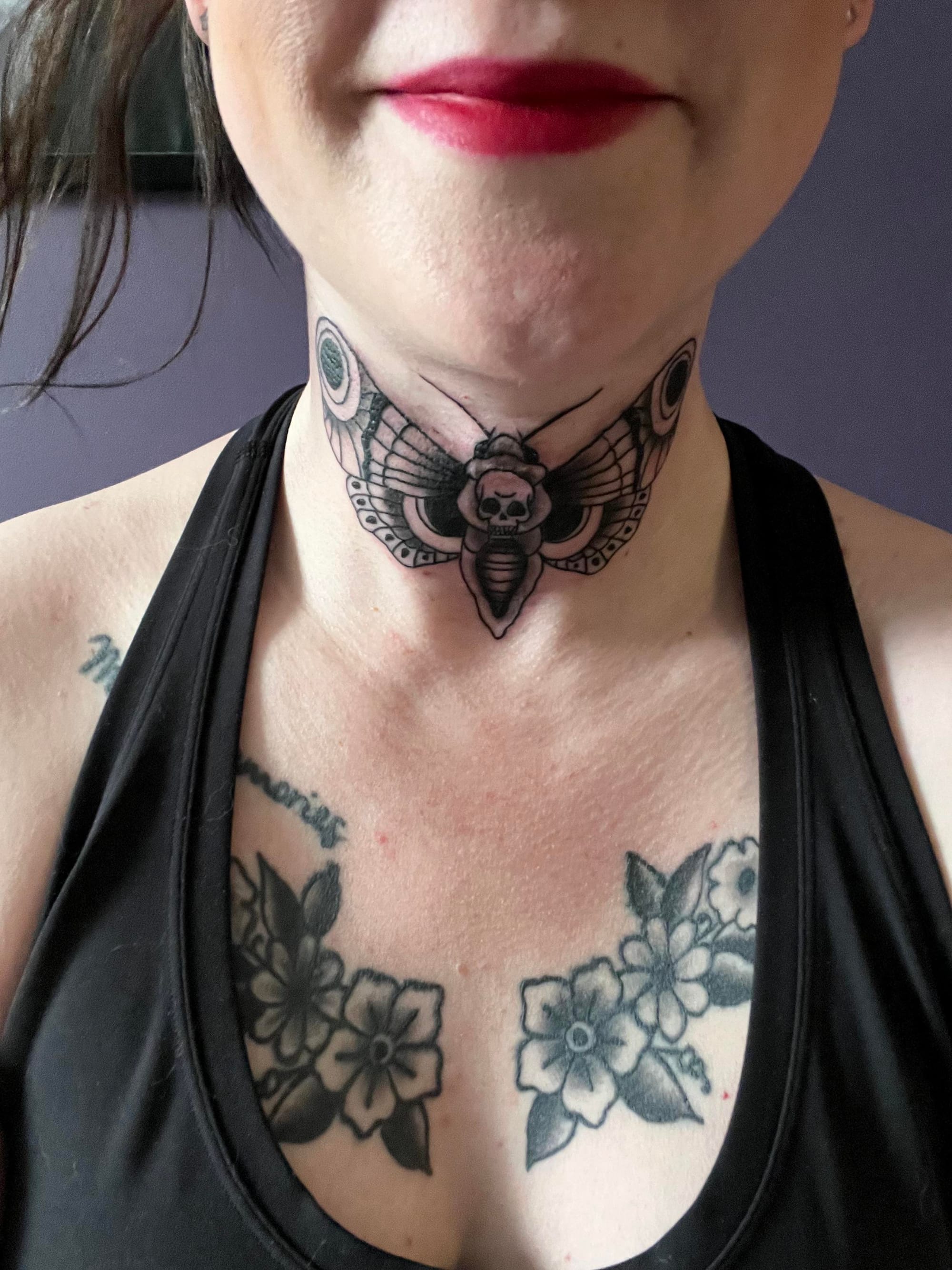 A hauntingly beautiful death's head hawkmoth spreads its wings across this throat placement, creating an incredibly bold statement piece that commands attention. The skull motif within the moth's body adds layers of gothic symbolism, while delicate floral tattoos on the chest create a stunning contrast between dark and light themes. 🦋💀