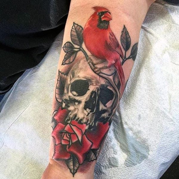 A striking juxtaposition of life and death unfolds across this forearm, where a vibrant red cardinal perches delicately above a weathered skull adorned with deep crimson roses. The masterful shading and realistic detail work creates a haunting beauty that speaks to themes of mortality, remembrance, and the eternal cycle of nature. This piece transforms traditional memento mori imagery into something unexpectedly hopeful through its brilliant pops of red against the monochromatic skull work.