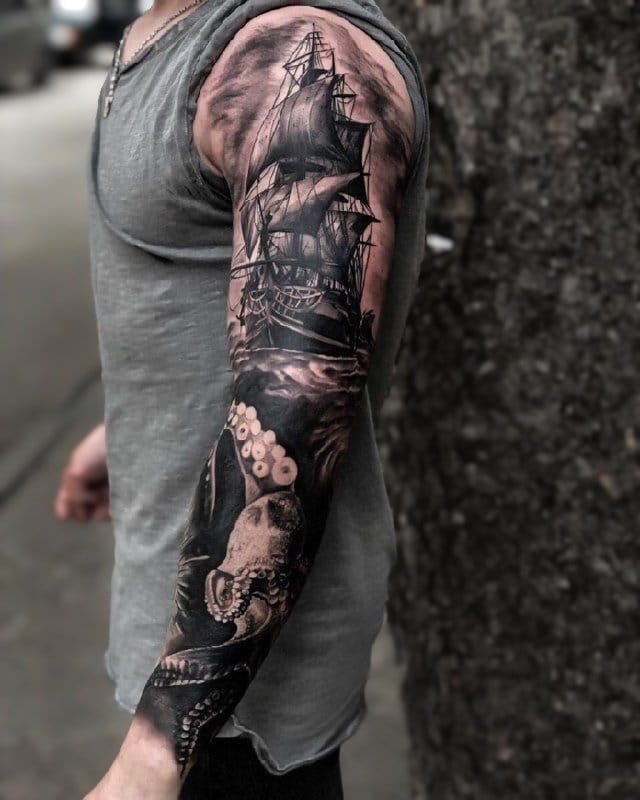 A masterful maritime saga unfolds across this full sleeve, where a majestic sailing ship battles through stormy seas while a colossal octopus emerges from the depths below. The incredible depth and shading work creates an almost three-dimensional effect, transforming the entire arm into a living piece of nautical folklore ⚓ The contrast between the billowing sails above and writhing tentacles below tells an epic tale of man versus nature in stunning black and grey realism.