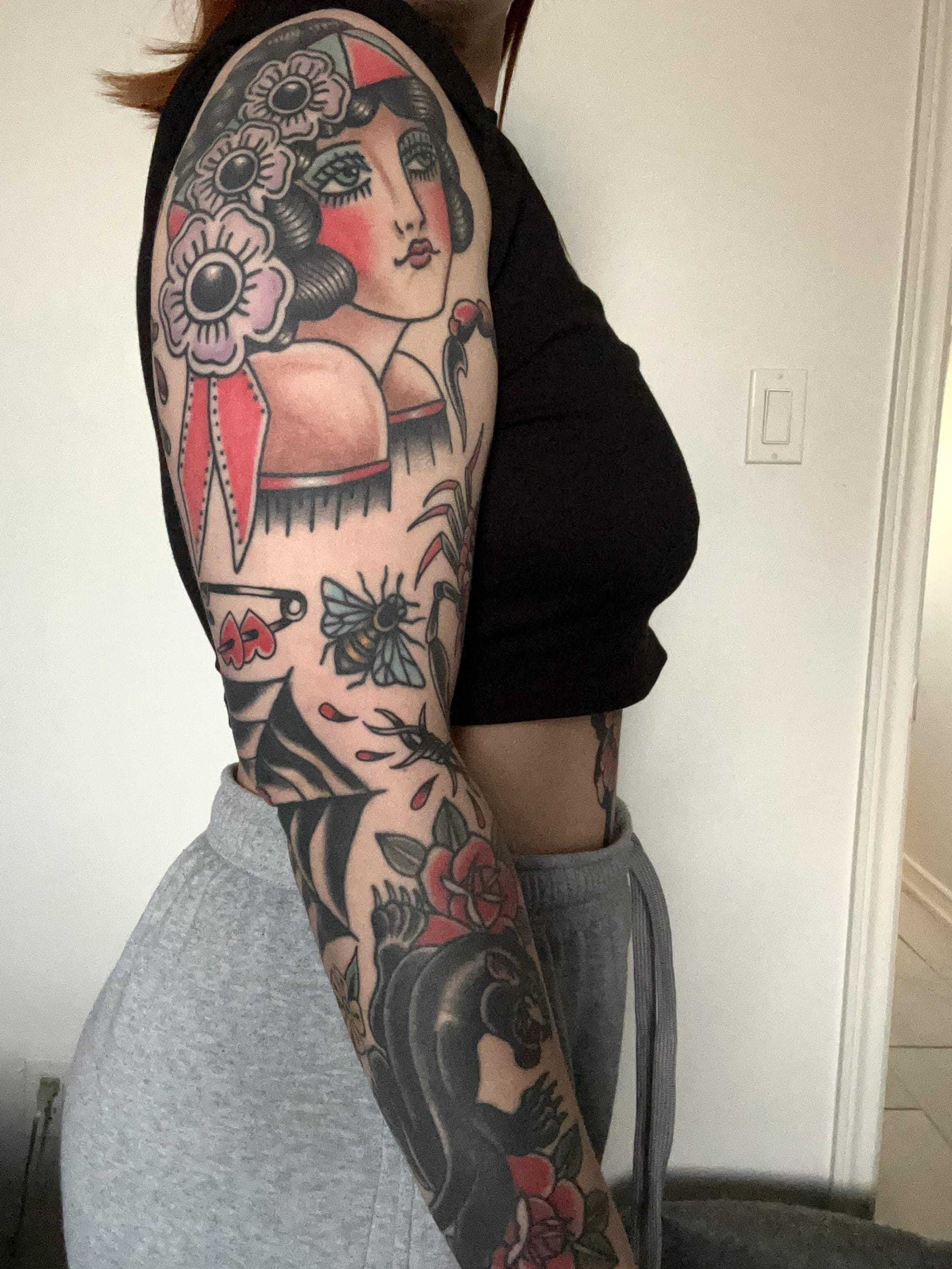 Bold traditional American style meets feminine mystique in this stunning full sleeve masterpiece. The artist's expert use of classic red, black, and green creates perfect harmony between the pin-up girl portrait, decorative florals, and nature elements that flow seamlessly from shoulder to wrist. Every inch tells a story while maintaining the timeless appeal of old school tattooing techniques. 🌹