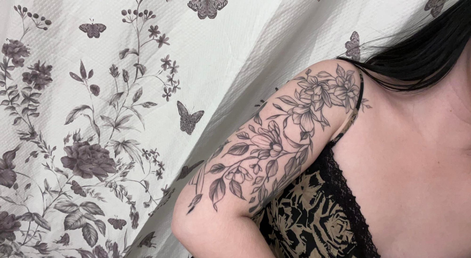 Delicate line work transforms this forearm into a botanical paradise, where intricate florals cascade in perfect harmony with the natural curve of the arm. The fine black ink creates stunning depth and movement, making each petal and leaf appear to dance across the skin 🌿 Notice how the design perfectly complements the floral wallpaper in the background - a dreamy match made in tattoo heaven!