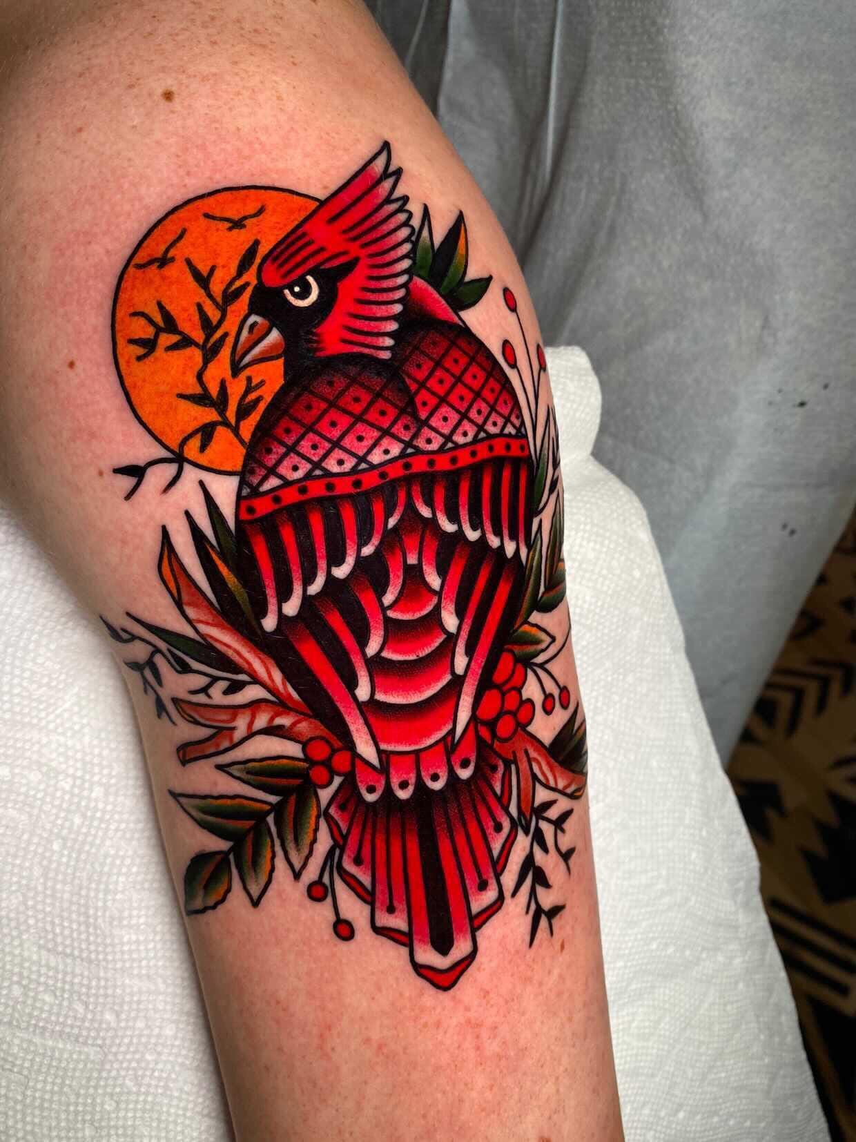 Bold traditional artistry takes flight in this stunning cardinal tattoo that perfectly captures the classic American style with its rich reds, deep blacks, and clean line work. The geometric patterns adorning the bird's chest create beautiful contrast against the organic foliage and setting sun backdrop, while the detailed feathering showcases the artist's technical skill. Positioned on the forearm, this piece commands attention with its vibrant color palette and timeless traditional tattoo aesthetic. 🔥