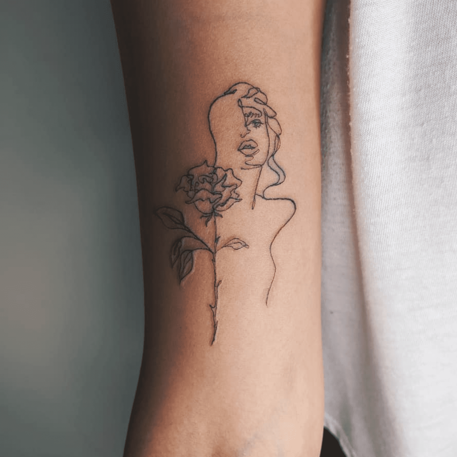 Delicate line work transforms skin into canvas with this minimalist masterpiece featuring a contemplative woman's profile intertwined with a blooming rose. The fine-line technique creates an almost sketch-like quality that feels both intimate and artistic, perfectly suited to the forearm's natural curve. There's something beautifully symbolic about the way the flower seems to grow from her essence, suggesting themes of inner beauty and personal growth 🌹