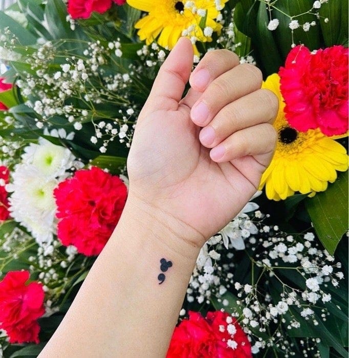 Delicate symbolism blooms on this wrist in the form of a minimalist semicolon tattoo, perfectly positioned against a backdrop of vibrant flowers. The subtle black ink design serves as a powerful reminder that life's story continues, while its placement allows for both meaningful visibility and easy concealment when desired. 🌸
