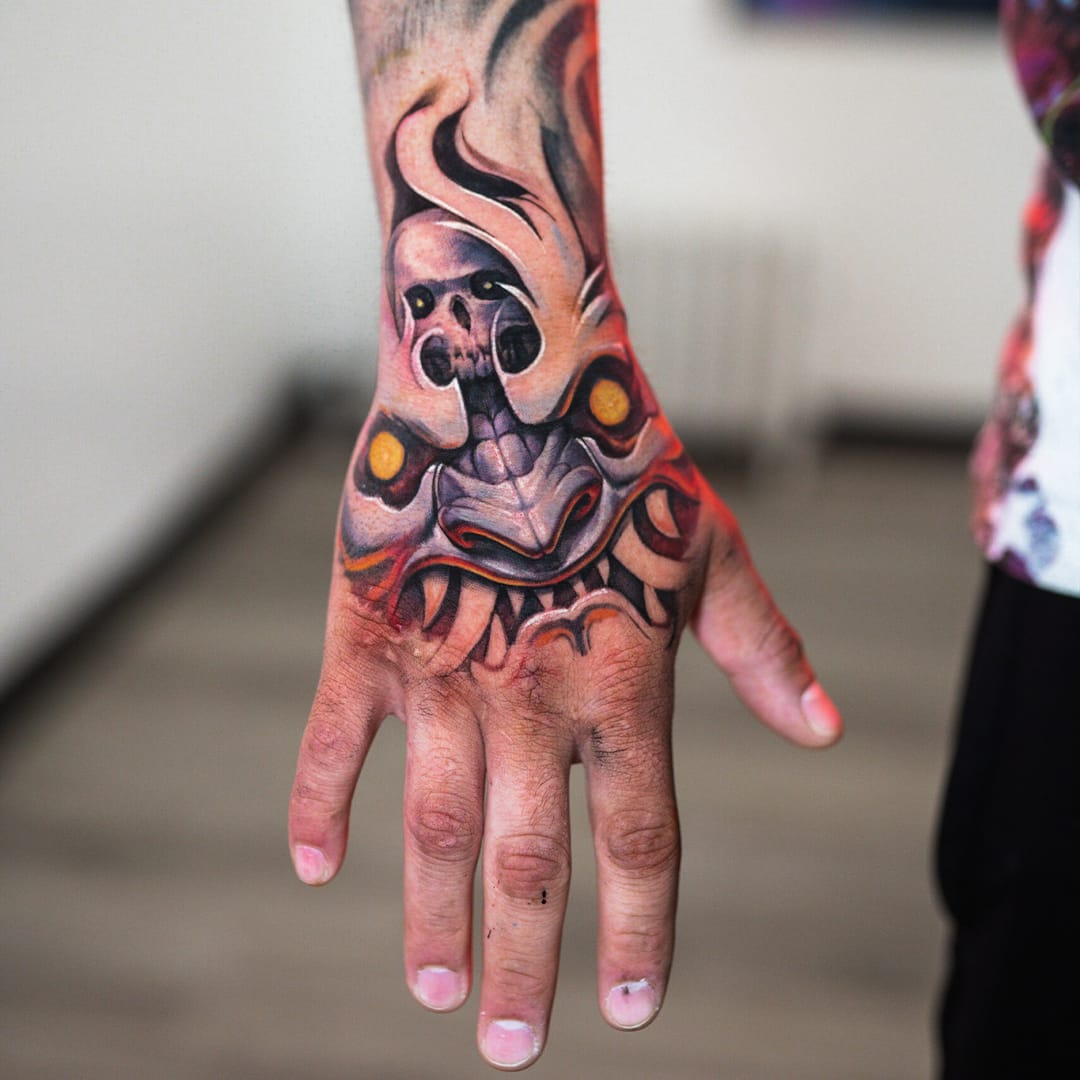 Demons and mythology collide in this powerfully executed hand tattoo, where a fierce oni mask commands attention with its menacing grin and hypnotic golden eyes. The artist's masterful use of traditional Japanese color palettes and bold linework transforms this everyday canvas into a statement piece that's impossible to ignore 👹