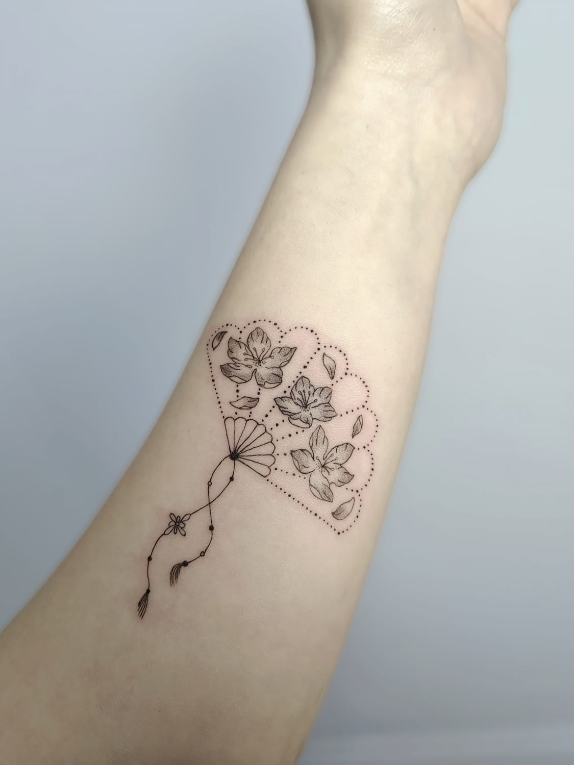 Delicate linework transforms this forearm into a botanical dreamscape, where three intricate flowers float within a cloud-like dotted border. The whimsical dangling elements below add movement and grace, creating a design that feels both grounded and ethereal ✨ Perfect placement for showcasing the fine detail work and organic flow of this nature-inspired piece.