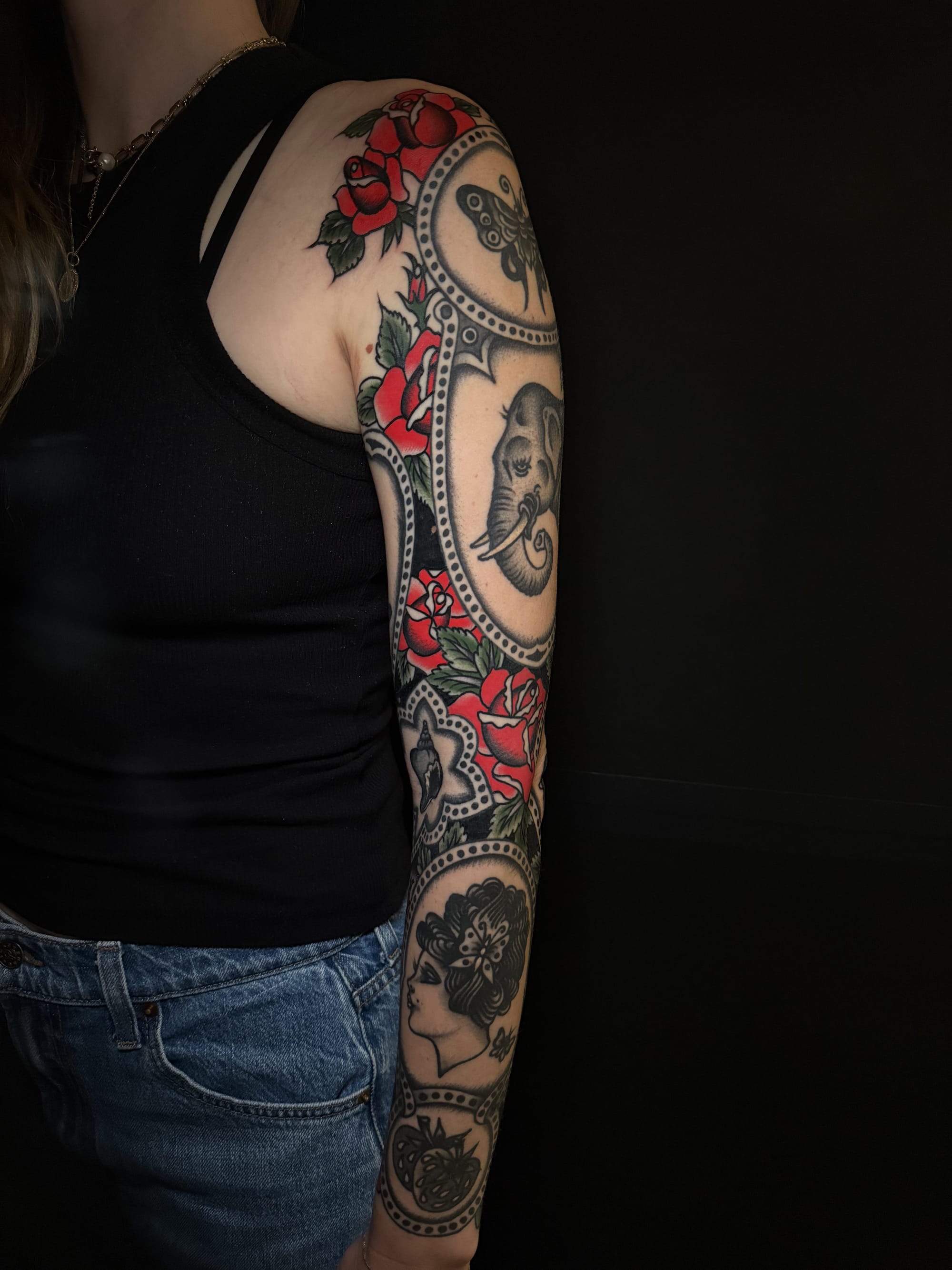 Cascading down this full sleeve like an enchanted garden, vibrant red roses weave through ornate decorative bands that frame mysterious portraits and intricate imagery. The masterful composition creates a stunning visual flow that transforms the entire arm into a living canvas of traditional tattoo artistry. Each circular vignette tells its own story while contributing to an absolutely breathtaking cohesive design 🌹