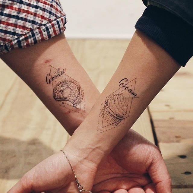 Sweet treats become eternal symbols of connection in this adorable couples' tattoo featuring matching cookie and cupcake designs on inner forearms. The delicate linework and geometric framing elevate these playful dessert motifs into sophisticated body art that celebrates a shared love of all things sugary. When their arms intertwine, these complementary tattoos create the perfect recipe for romance 🧁