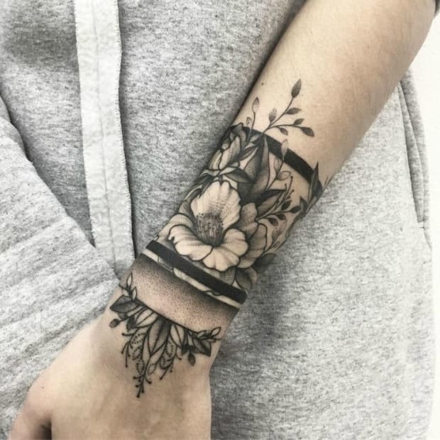 Botanical artistry meets contemporary design in this striking forearm piece, where delicate florals flow seamlessly around what appears to be a decorative band or bracelet element. The contrast between the organic curves of leaves and petals against the geometric structure creates a captivating visual balance that's both feminine and bold. 🌿 The way the design wraps around the wrist and extends up the arm transforms the entire area into a living canvas of nature-inspired beauty.