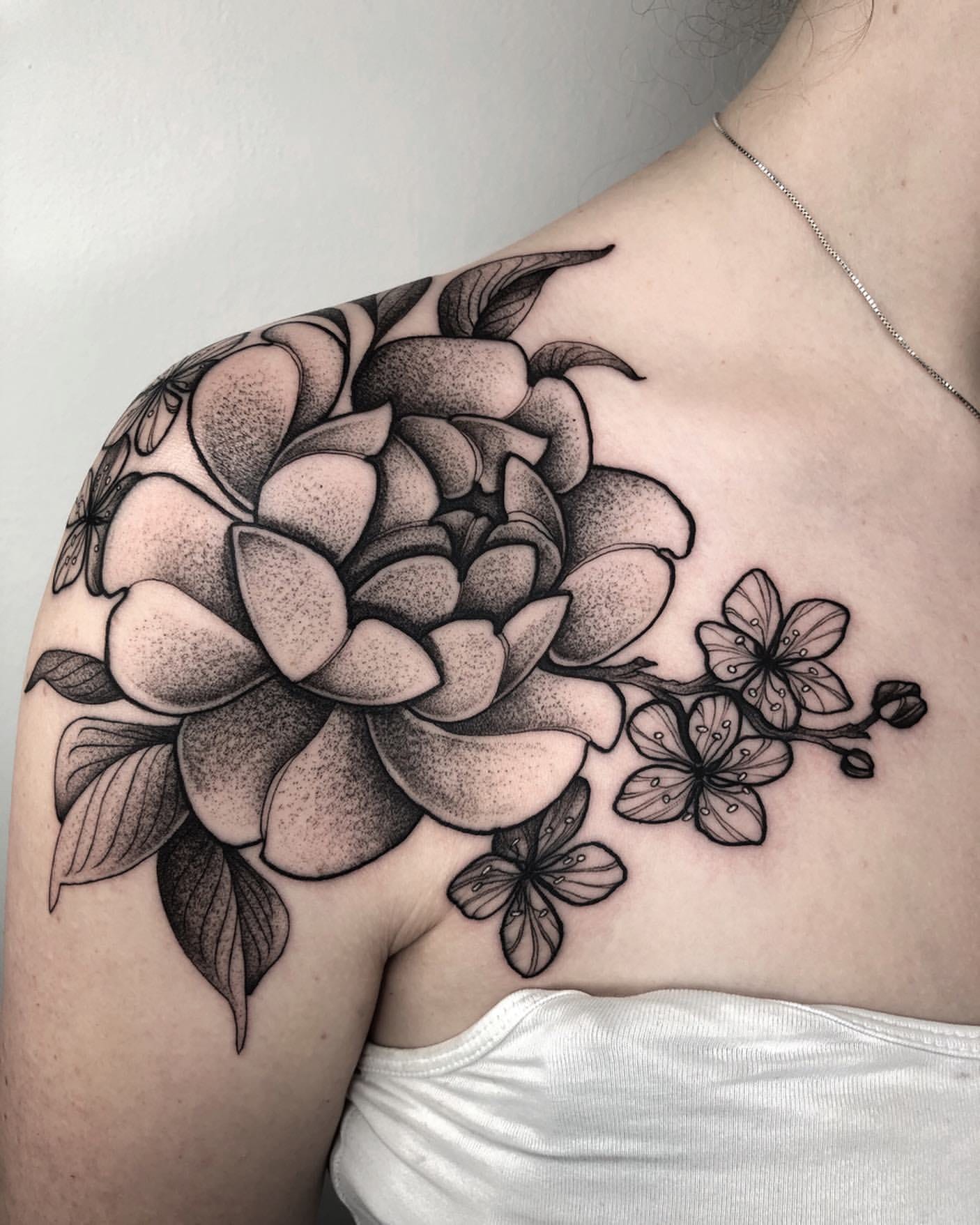 Botanical elegance blooms across this shoulder blade in the most stunning blackwork floral composition 🌸 The artist's masterful dotwork technique creates incredible depth and texture, making each peony petal appear almost three-dimensional against the skin. Notice how the smaller blossoms cascade gracefully toward the collarbone, creating a natural flow that perfectly complements the body's curves.