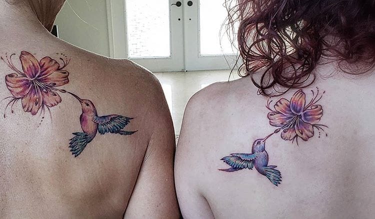 Delicate hummingbirds dance toward blooming hibiscus flowers in these perfectly mirrored shoulder blade tattoos that celebrate the beauty of shared experiences. The soft watercolor technique brings dreamy pastels to life, creating an enchanting garden scene that flows naturally with the curve of each shoulder 🌺 What makes this design truly special is how the birds appear to be flying toward each other when the two people stand together, symbolizing connection and harmony.
