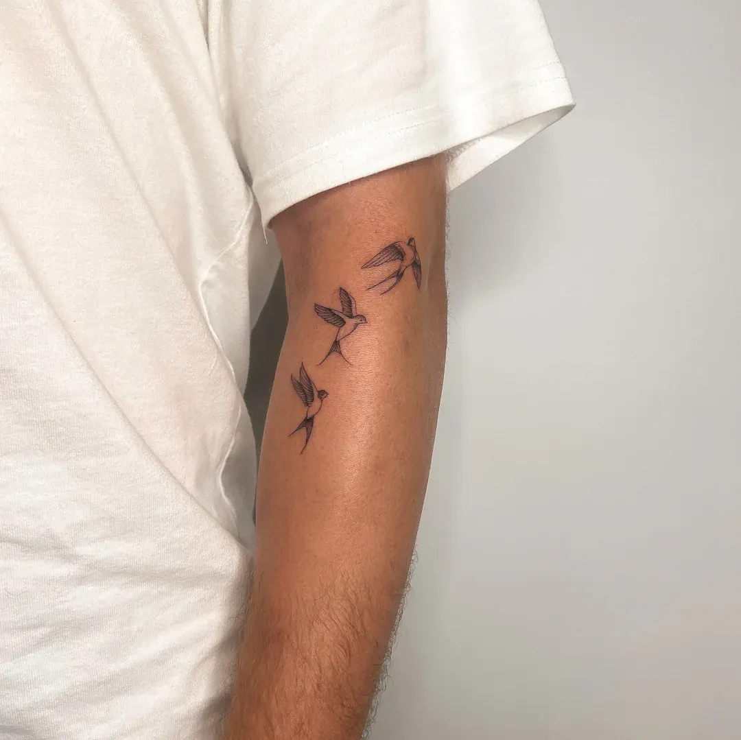 Three delicate hummingbirds take flight along this forearm, their fine-line silhouettes capturing the essence of freedom and grace 🕊️ The minimalist black ink work creates a stunning visual flow that follows the natural curve of the arm, making it appear as though these tiny creatures are genuinely soaring across the skin. Perfect placement for a design that celebrates life's fleeting beautiful moments.