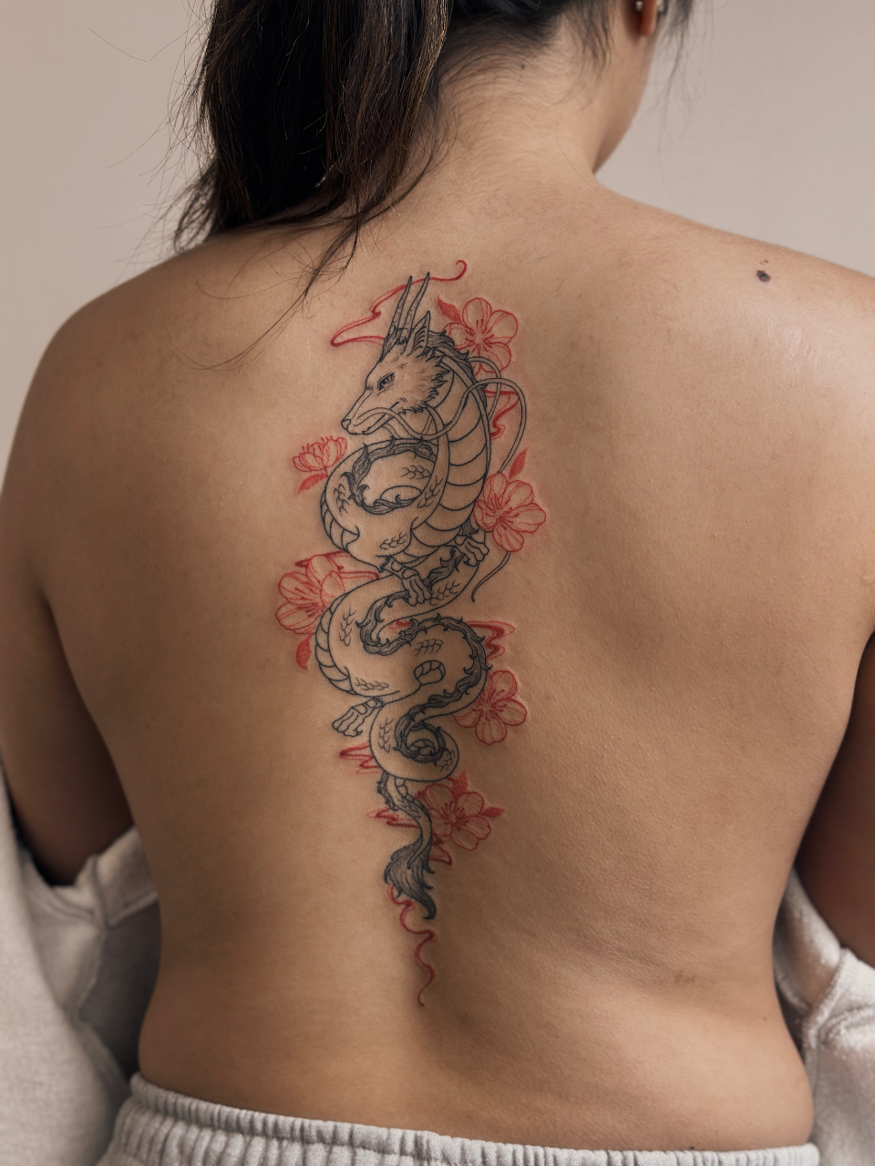 Gracefully winding down the spine, this ethereal dragon design combines delicate line work with vibrant cherry blossoms in a stunning display of Eastern artistry. The serpentine creature's flowing form perfectly follows the natural curve of the back, while soft pink florals add feminine touches to the powerful mythological symbol. A masterpiece of placement that transforms the entire back into a canvas of ancient folklore and botanical beauty.