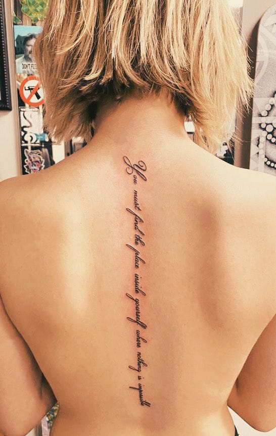 Cascading down the spine like a whispered secret, this delicate script tattoo flows from neck to lower back with effortless grace. The cursive lettering creates a stunning vertical statement piece, while the small bow detail at the top adds a touch of feminine charm that perfectly complements the flowing text below. ✨