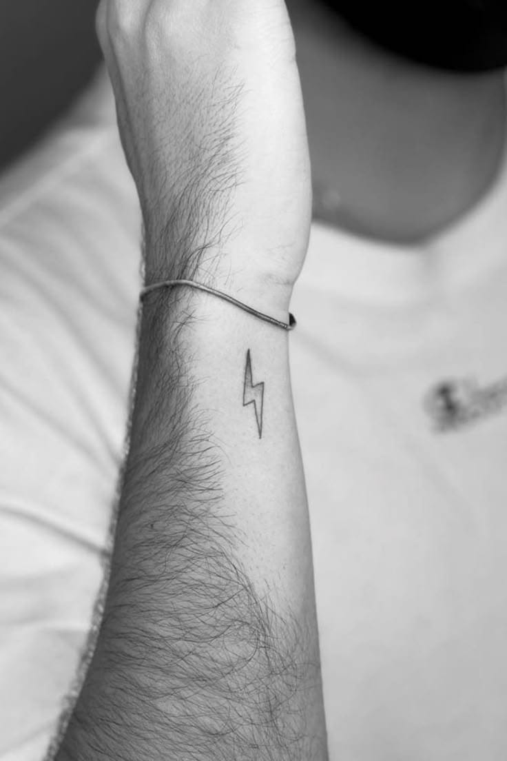 Bold minimalism meets symbolic power in this striking lightning bolt tattoo that commands attention on the forearm. The clean black linework creates a sharp contrast against the skin, while its strategic placement just below a simple bracelet adds an edgy accent to everyday style ⚡ Perfect for those who want to carry a reminder of their inner strength and electric energy wherever they go.