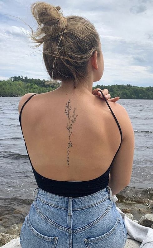 Delicate botanical artistry flows down this woman's spine like nature's own calligraphy, creating an enchanting vertical garden against sun-kissed skin. The minimalist line work and graceful placement transform her back into a living canvas, perfectly complemented by the serene lakeside setting 🌿