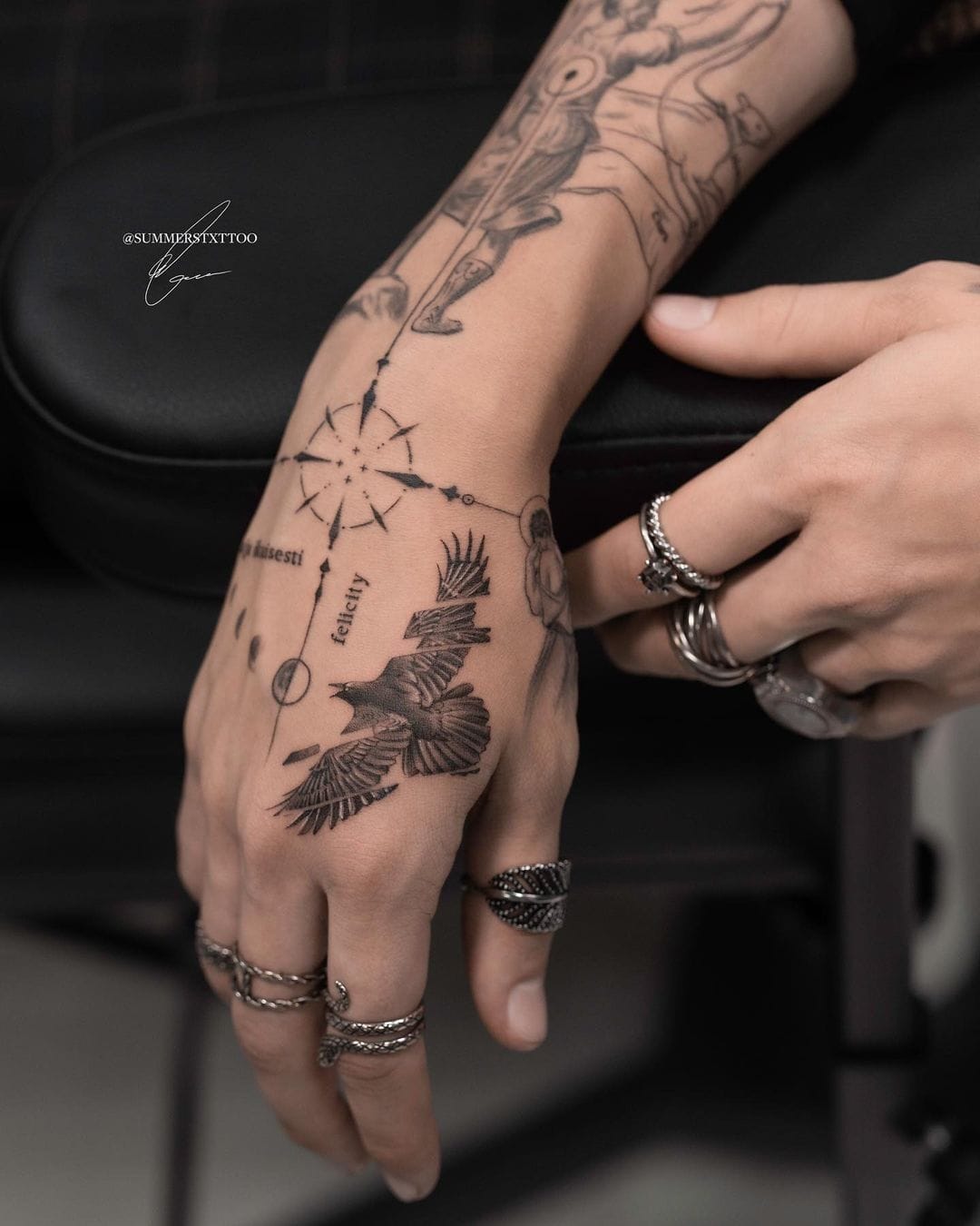 Wanderlust meets artistry in this stunning forearm and hand tattoo featuring a soaring eagle, compass rose, and decorative elements that flow seamlessly from wrist to knuckles. The intricate linework and strategic placement create a cohesive story of freedom and direction, while those statement silver rings add the perfect finishing touch to this bold body art statement. 🧭