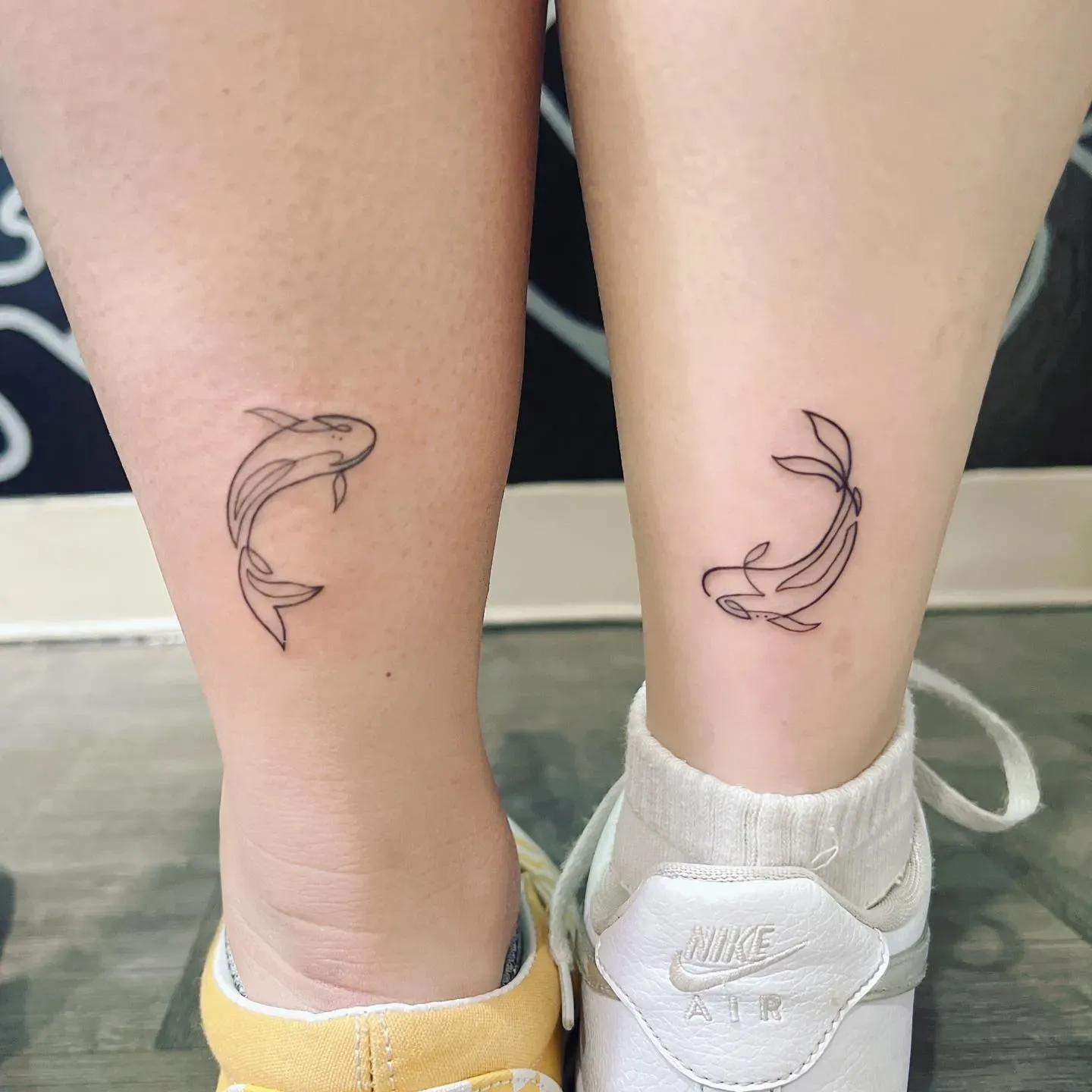Minimalist line art transforms these matching ankle tattoos into a stunning display of duality - two koi fish swimming in opposite directions, creating perfect visual harmony across both legs. The delicate black outlines capture the graceful movement of these symbolic creatures, representing balance, perseverance, and the beautiful flow of life's journey 🐟 Positioned just above white sneakers, these complementary designs prove that sometimes the most powerful tattoos speak through simplicity rather than bold statements.