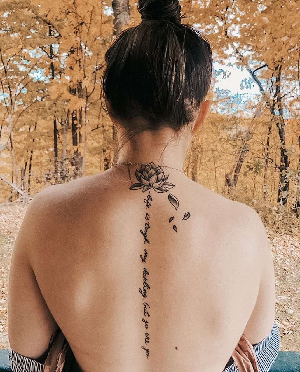 Delicate script cascades down the spine like fallen petals, crowned by an intricate lotus bloom that seems to float at the base of the neck. The graceful vertical text creates an elegant pathway from shoulder blade to lower back, while scattered leaf motifs add organic movement to this stunning botanical design. Pure poetry written on skin against autumn's golden backdrop. 🍂